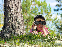 Das Foto zeigt eine Schülerin die sich neben einem Baum im hohen Gras versteckt und mit dem Fernglas die Wildtiere rund um Spital am Pyhrn beobachtet.