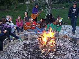 Das Foto zeigt unsere SchülerInnen beim Lagerfeuer. Einige SchülerInnen und LehrerInnen spielen mit der Gitarre Lieder und schaffen damit noch mehr Lagerfeuerromantik.