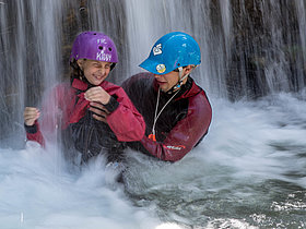 Das Bild zeigt eine Schülerin und einen unserer geschulten Guides mitten in einem Wasserfall beim Jugendschluchtenwandern.