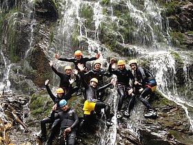 Das Foto zeigt unsere Schüler und Schülerinnen beim Canyoning. Die Gruppe posiert hier für ein cooles Erinnerungsfoto. Während einer unserer Guides mit der gelben Hose den Felsen hoch zu klettern scheint, machen es sich die restlichen Kinder und Guides auf dem Felsen bequem. im Hintergrund prasselt das Wasser eines Wasserfalls herunter