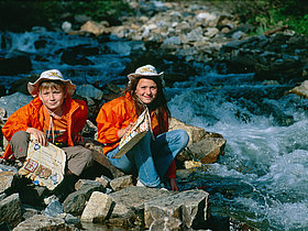 Das Foto zeigt zwei SchülerInnen mit oranger Jacke und Cowboy Hut am Ufer des Kremsbaches. Beide halten ihre Schatzkarte in der Hand und das Mädchen hält die Hand ins glasklare Wasser des Kremsbaches