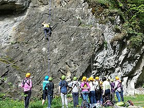 Das Bild zeigt Kinder am Kletterfelsen beim Programm CAM Mölltal 5 Tage. Ein Kletterer mit gelbem Shirt klettert gut gesichert an der Kante während die restlichen Kinder gespannt zusehen und sich aufs Klettern vorbereiten. 