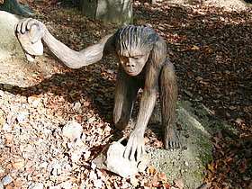 das Foto zeigt das lebensgroße Modell eines Urzeitmenschen mitten im Styrassic Park mit seinen unzähligen lebensgroßen Modellen verschiedener Saurier