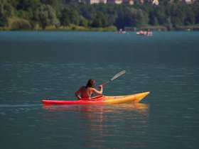 Das Foto zeigt den Naturteich im WAHAHA Paradise. Der Naturteich ist von Schilfgürteln, Steinen und Sträuchern umgeben. im Hintergrund sehen wir links den Badesteg und in der Bildmitte die Saunalandschaft 